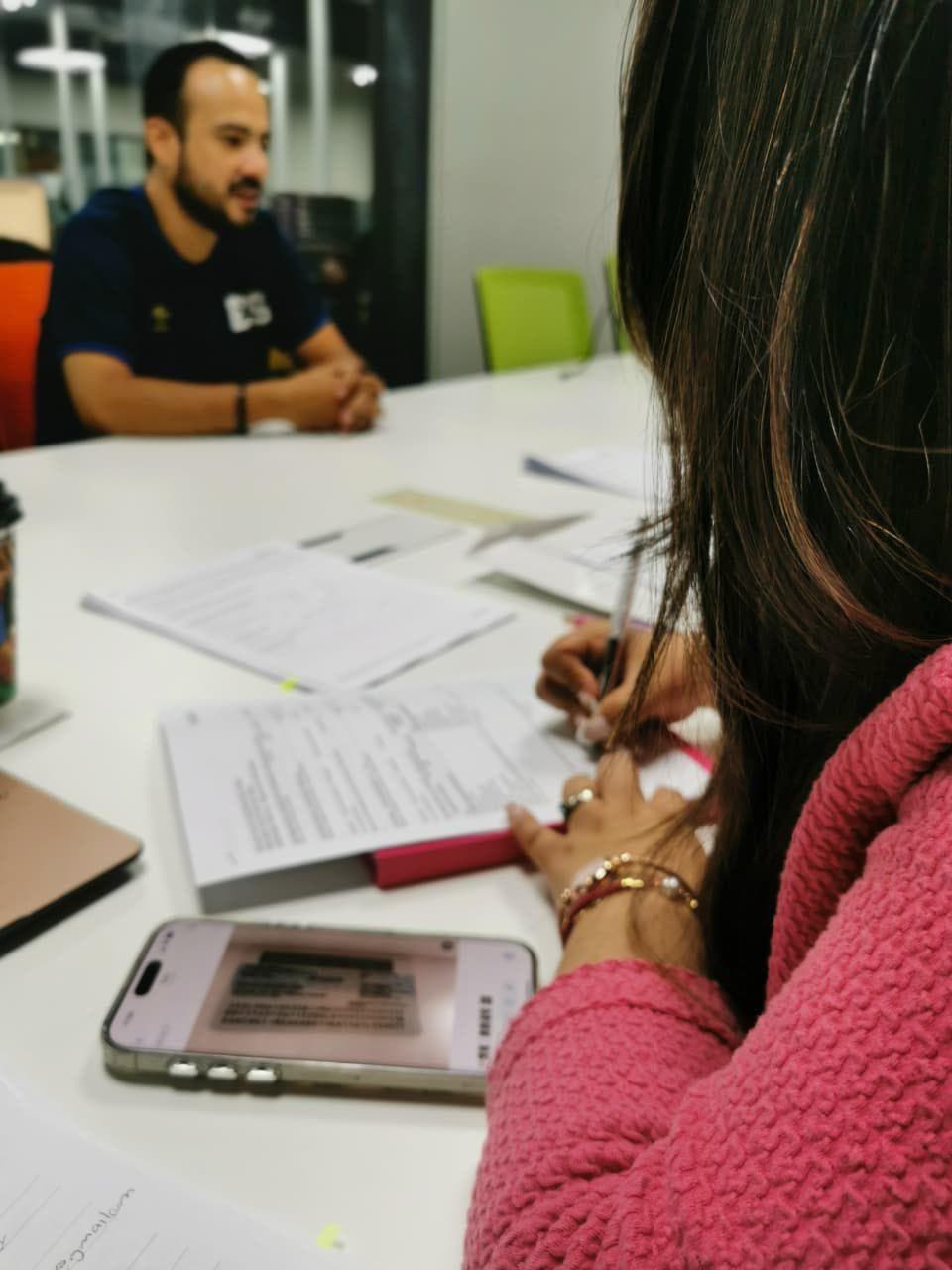Mujer con suéter rosa firma documentos en una mesa frente a un hombre desenfocado.