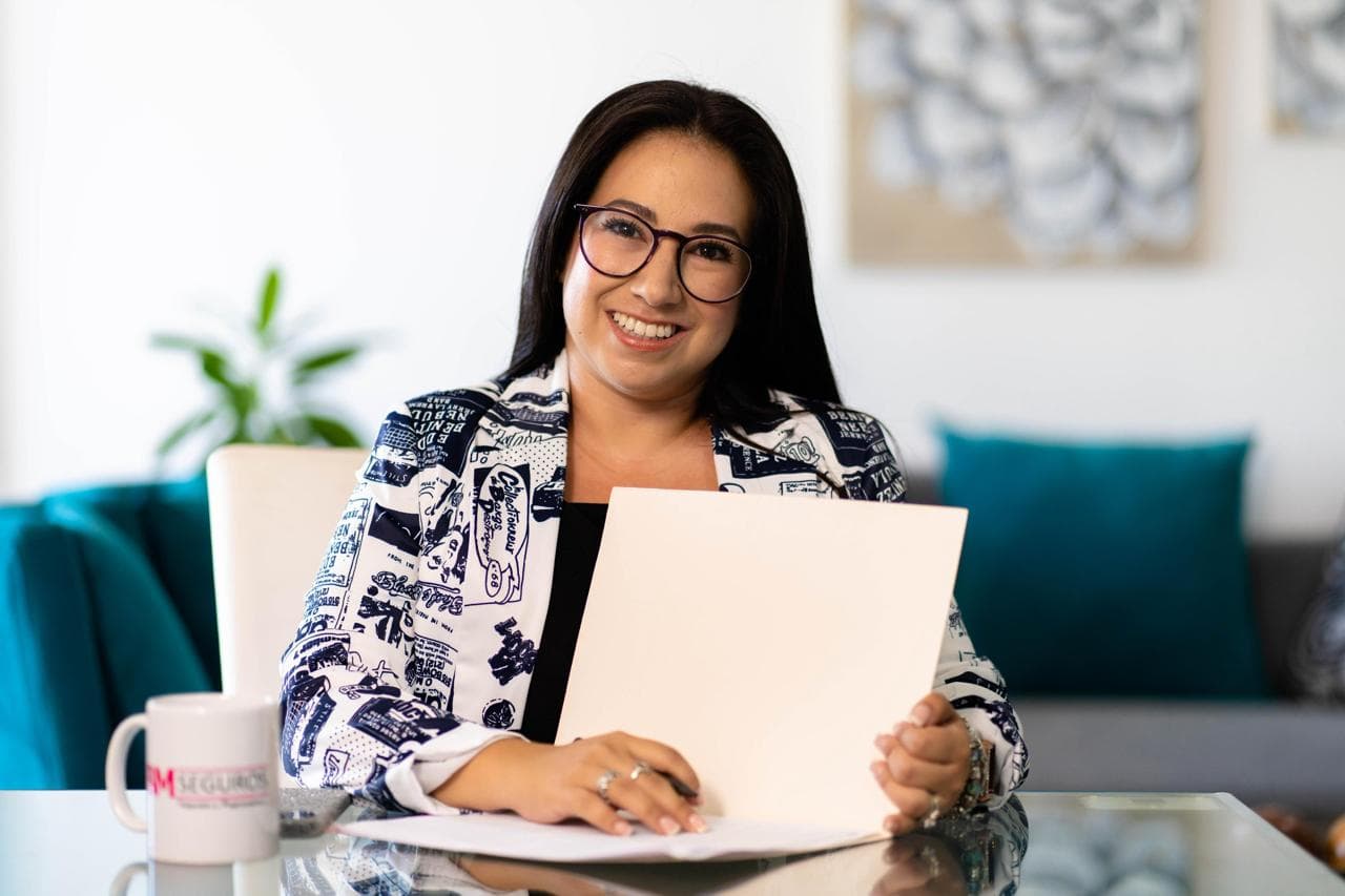 Mujer sonriente con gafas y chaqueta estampada sosteniendo una carpeta blanca en un escritorio.